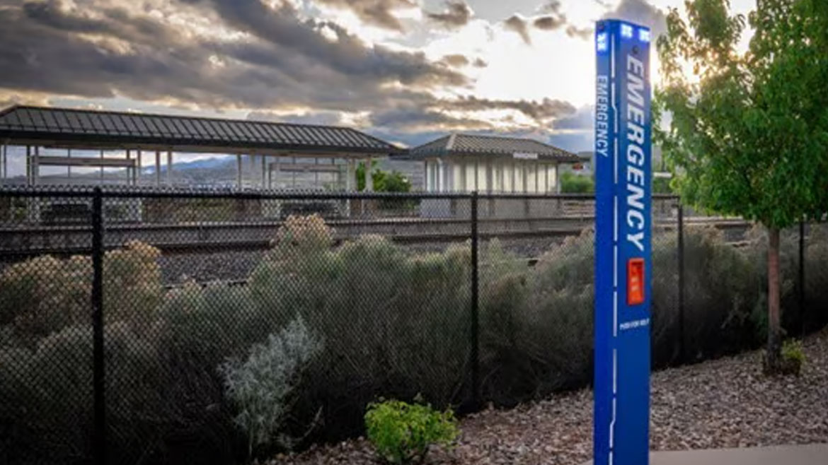 Blue emergency call box at a train station with tracks, a fence, and a cloudy sky.